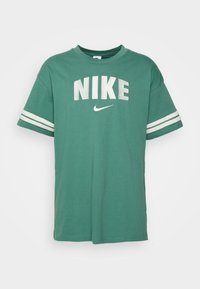 Grön t-shirt med kort ärm, gjord av bomull och med rund halsringning, som har "NIKE" i stora vita bokstäver och vita ränder på ärmarna.