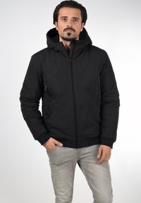 Veste noire zippée avec capuche, dotée de poches latérales, de poignets côtelés et d'un léger éclat. Texture lisse et design minimaliste.