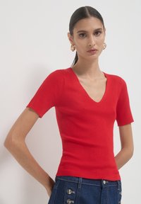 Junge Frau mit dunklem Haar im Pferdeschwanz, die goldene Creolenohrringe, ein rotes, geripptes Kurzarm-V-Top und dunkelblaue, geknöpfte Jeans trägt.