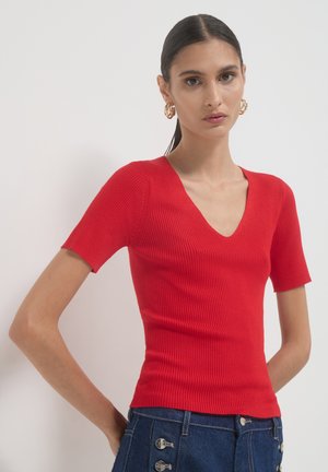 Junge Frau mit dunklem Haar im Pferdeschwanz, die goldene Creolenohrringe, ein rotes, geripptes Kurzarm-V-Top und dunkelblaue, geknöpfte Jeans trägt.