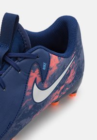 Náutro modré futbalové kopačky s textúrovaným zvrškom, ktorý má ružové a oranžové abstraktné vzory, strieborný Nike znak a oranžové trakčné kolíky.