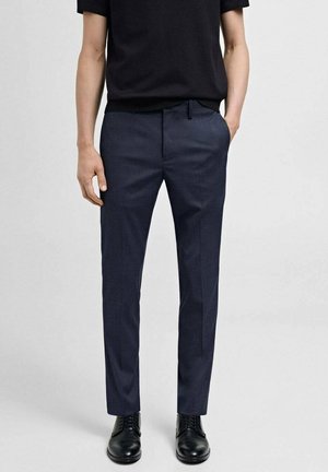 Mann trägt marineblaue formelle Hose, schwarze Lederschuhe und ein schwarzes kurzärmeliges Hemd, steht mit einer Hand in der Tasche.