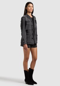 Vrouw staand, draagt een donkergrijze blouse met knopen en meerdere zakken, bijpassende korte broek en zwarte laarzen tot halverwege de kuit, tegen een lichte achtergrond.