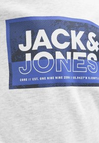 Maglietta grigia chiara con una grafica rettangolare blu audace che comprende il testo "JACK & JONES" e coordinate, con uno sfondo texturizzato.
