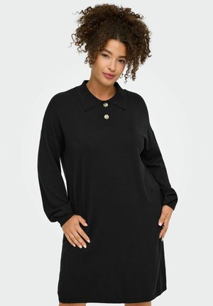 Femme aux cheveux bouclés portant une longue robe noire à col avec deux boutons décoratifs, posant avec les mains sur les hanches devant un fond uni.