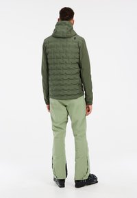 Doudoune verte avec une capuche et un devant texturé ; associée à un pantalon vert clair et des bottes de ski noires ; branding minimal sur la doudoune.