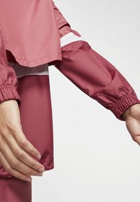 Veste imperméable rose avec poignets élastiques, un design à couches superposées, et des bandes accentuées blanches sur les manches, présentant une texture lisse et brillante.