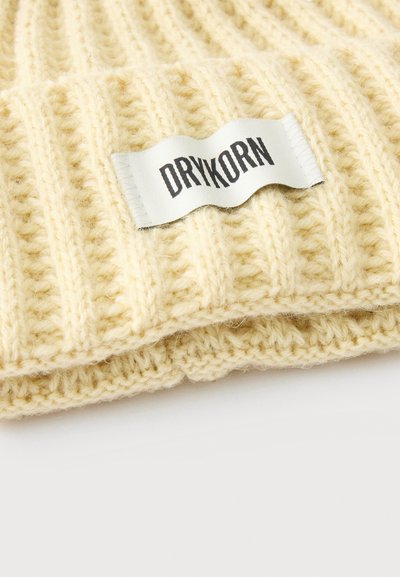 Tissu tricoté côtelé de couleur crème avec une étiquette rectangulaire blanche affichant le nom de la marque "DRYKORN" en lettres noires.