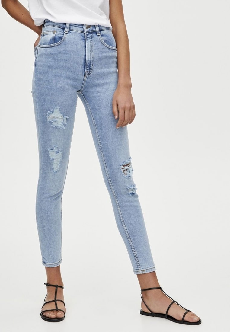 Jeans de tiro alto de color azul claro con detalles desgatados, incluyendo roturas y bordes deshilachados, combinados con sandalias negras de tiras.
