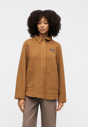 Frau trägt eine braune, strukturierte Fleecejacke mit Kragen und vorderen Taschen und steht vor einem schlichten hellen Hintergrund.