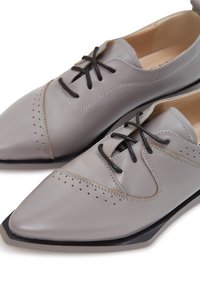 Chaussures en cuir gris avec un bout pointu, dotées de lacets noirs et de perforations décoratives. Texture lisse avec une semelle noire contrastante.