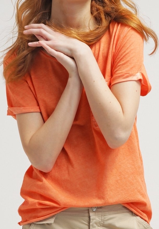 Osoba s červenými vlasy, oblečená v oranžovém tričku a béžových kalhotách, se sepjatýma rukama u krku na jednobarevném pozadí.