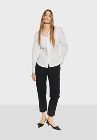 Camicia bianca a maniche lunghe con dettagli a bottoni, abbinata a pantaloni neri a vita alta e tacchi neri a punta con dettaglio a fiocco.