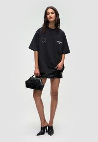 T-shirt noir oversize avec un logo imprimé, associé à une mini-jupe en simili cuir noir brillant et un sac à main noir texturé. Mules noires à bouts pointus.