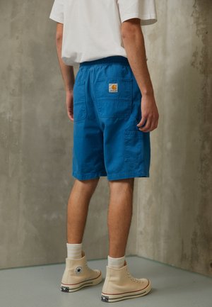 Shorts en coton bleus avec une coupe décontractée, dotés de grandes poches latérales et d'une étiquette tissée à l'arrière. Portés avec des baskets montantes beige.