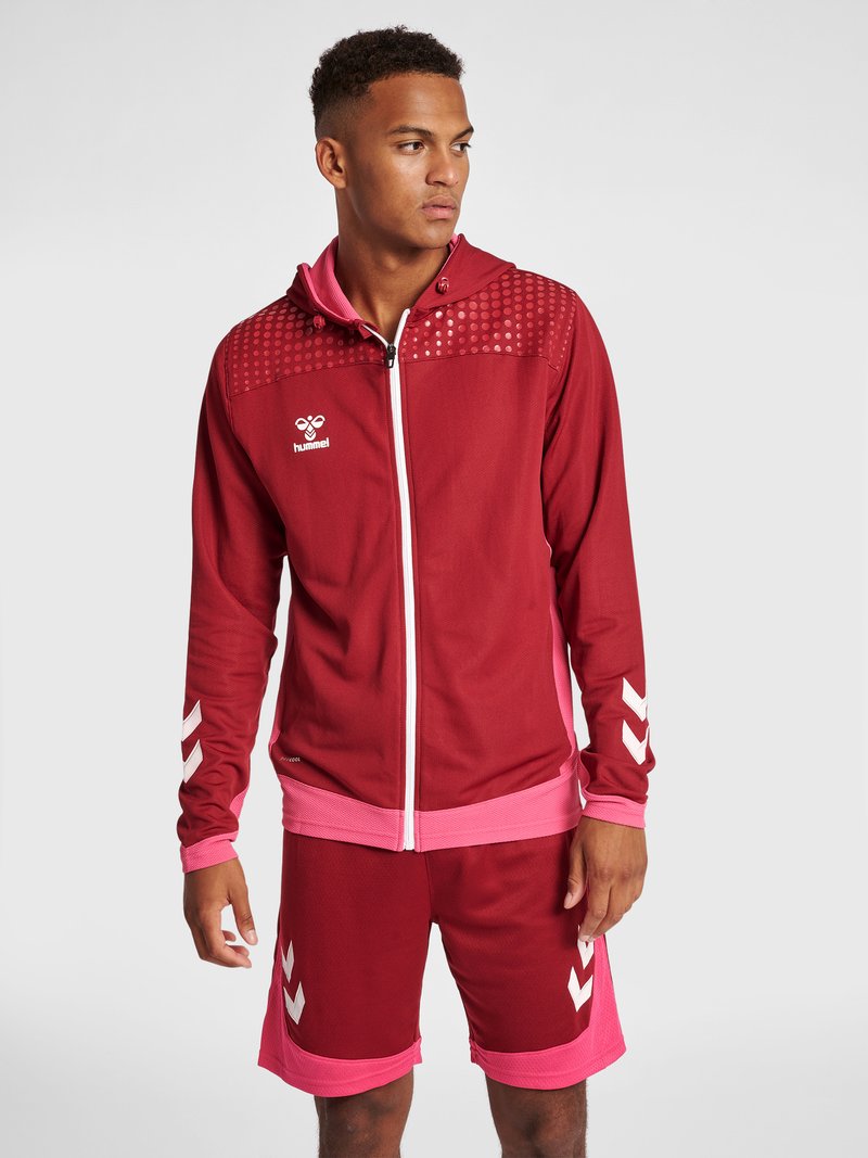 Man die een rood en roze sportjack en bijpassende korte broek draagt, staande tegen een effen lichte achtergrond, kijkt opzij.