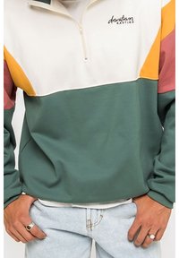 Pullover a blocchi di colore in verde, bianco, arancione e rosa. Presenta un collo con mezza zip e un logo sul petto. Tessuto morbido, vestibilità comoda.