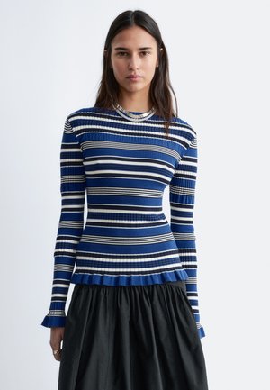 Woman wearing a fitted blue, white, and black striped long-sleeve sweater with ruffled hem and black skirt, standing against a plain background.