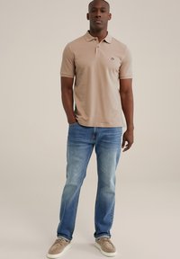 Beige pikétröja med kort ärm och krage, försedd med en liten logotyp på bröstet. Bärs tillsammans med ljusblå denimjeans och beige sneakers.