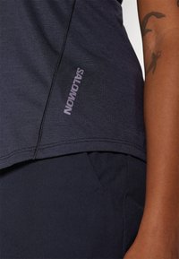 Mørkegrå idrettstopp med flate sømmer og en liten "Salomon"-logo. Stoffet har en jevn tekstur, designet for komfort og ytelse.