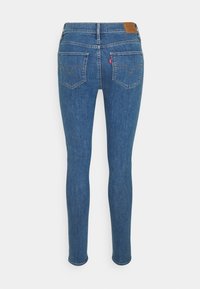 Jean slim bleu avec poches arrière, passants de ceinture et petite pièce en cuir sur la ceinture.