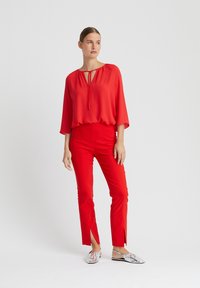 Rote, fließende Bluse mit einem Schlüsselloch-Ausschnitt, kombiniert mit figurbetonten roten Hosen mit seitlichen Schlitzen. Silberne, strukturierte Slipper.