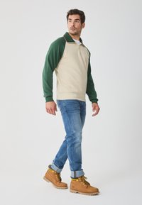 Suéter beige con mangas y cuello verdes, con un diseño de media cremallera. Combinado con jeans azules y botas marrones con cordones amarillos.