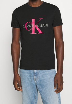 Personne portant un t-shirt noir Calvin Klein Jeans avec un grand logo "cK" rose et un jean en denim bleu clair sur un fond uni.