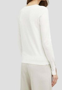 Vit långärmad tröja med rund halsringning och ribbad fåll, tillverkad av ett slätt, lätt tyg; bärs över ljusbeiga byxor.