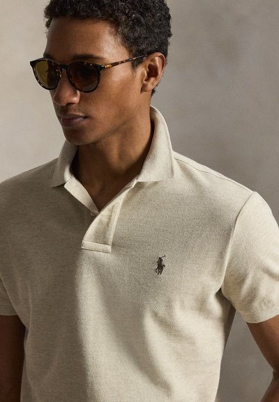 Man draagt een zonnebril met schildpadmotief en een beige poloshirt met een klein geborduurd logo op de borst, kijkt zijwaarts.