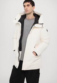 Parka blanche avec une doublure noire, dotée d'un col montant, d'une fermeture éclair, d'une grande capuche et de poches avant. Pull gris côtelé porté en dessous.