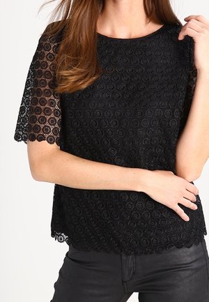 Zwart kanten top met korte mouwen en ronde patronen, een ronde halslijn en een gegolfde zoom. Gedragen over een zwarte broek.