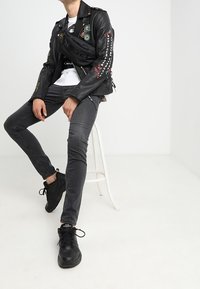 Chaqueta de biker de cuero negro con parches, jeans negros ajustados y zapatillas negras. Diseño robusto con cremalleras y tachuelas, sentado en un taburete blanco.