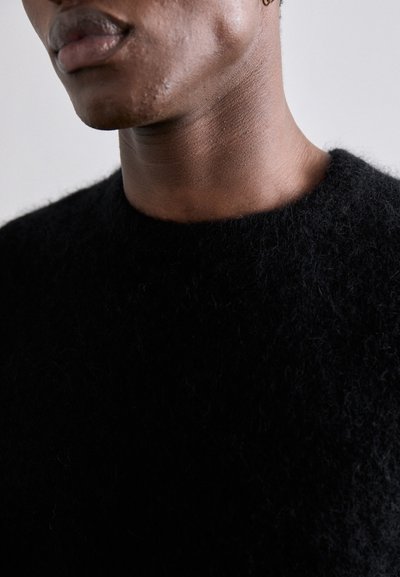 Suéter negro, peludo, con una textura suave, cuello alto y sin patrones visibles, llevado por una persona que muestra solo el cuello y parte de la cara.