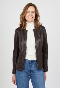 Braune Lederjacke mit rundem Kragen, tailliertem Design und Reißverschlusstaschen, getragen über einem weißen Rollkragenpullover und blauen Jeans.