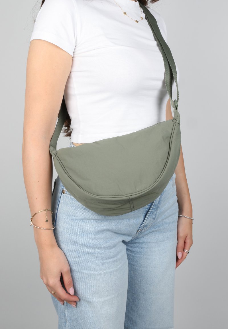 Donna che indossa jeans azzurri chiari e una maglietta bianca, con una borsa a tracolla verde oliva attraverso il petto e la spalla.