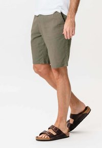 Mand iført olivengrønne shorts og brune sandaler med dobbelt spænde, stående med det ene ben let bøjet mod en hvid baggrund.