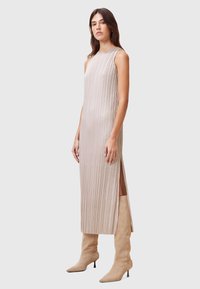 Abito lungo senza maniche di colore beige con pieghe verticali, fessure laterali e una texture liscia. Abbinato a stivali beige a punta con altezze fino al ginocchio.