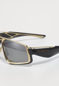 Černé a zlaté sluneční brýle Oakley s obdélníkovými tónovanými čočkami a nápisem "Prizm P" vytištěným v rohu čočky.