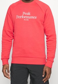 Rode sweatshirt met ribgebreide boorden en onderrand, voorzien van het "Peak Performance"-logo in het wit op de voorkant, gemaakt van zachte stof.