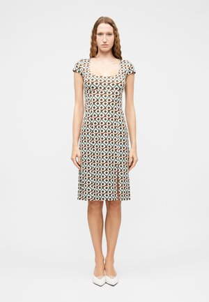 Kobieta z długimi falującymi włosami, ubrana w krótką, wzorzystą sukienkę do kolan z przednim rozporkiem i białe, spiczaste buty, stojąca prosto.