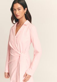 Rozā svītrains wrap tipa kleita ar garām piedurknēm, apkakli un sasienamu vidukli; izgatavota no gludas auduma ar vertikālām baltām svītrām.