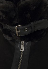 Svart konstläderkrage på en svart mockajacka, med en silverdragkedja och ett läderband med en metallspänne. Texturerad och smidig design.