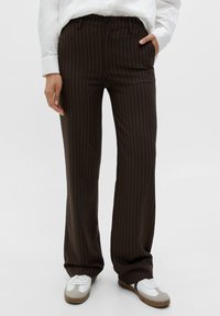 Bruna prickiga byxor i slätt tyg, med platt front, sidofickor och avslappnad passform, tillsammans med vita och grå sneakers.