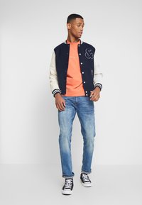 Marineblå varsityjakke med kremfargede ermer, en brodert logo, oransje genser under, lyseblå jeans og svarte sneakers med hvite detaljer.