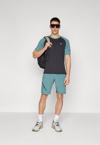Pánský sportovní outfit s černým a tyrkysovým tričkem s krátkým rukávem, tyrkysovými šortkami, bílými ponožkami, šedými teniskami a černým batohem.