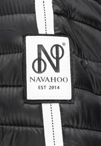 Tissu matelassé noir avec une étiquette de marque rectangulaire blanche portant l'inscription noire "NAVAHOO" et "EST 2014". Détails de design avec des rayures blanches verticales.