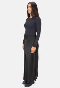 Schwarzes langärmliges Oberteil, kombiniert mit einem fließenden schwarzen Satin-Maxikleid, das eine elegante Silhouette und strukturierte Stoffdetails aufweist. Schwarze Schuhe runden den Look ab.