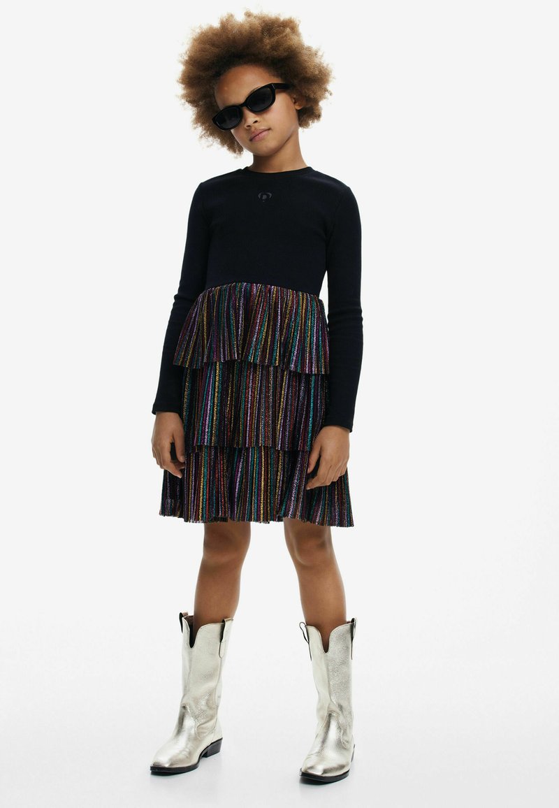 Top nero a maniche lunghe con una gonna a strati a righe multicolori. L'outfit è abbinato a stivali da cowboy argentati fino al ginocchio e occhiali da sole neri.