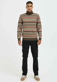 Pulover cu guler înalt, de culoare bej, cu modele geometrice intricate în nuanțe de maro, roșu și crem. Asortat cu pantaloni negri și adidași gri.